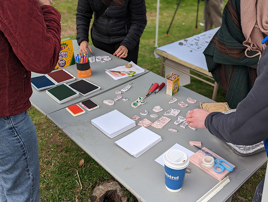 stamps are displayed with hands of participants, pieces of paper, crayons in a grassy outdoor space